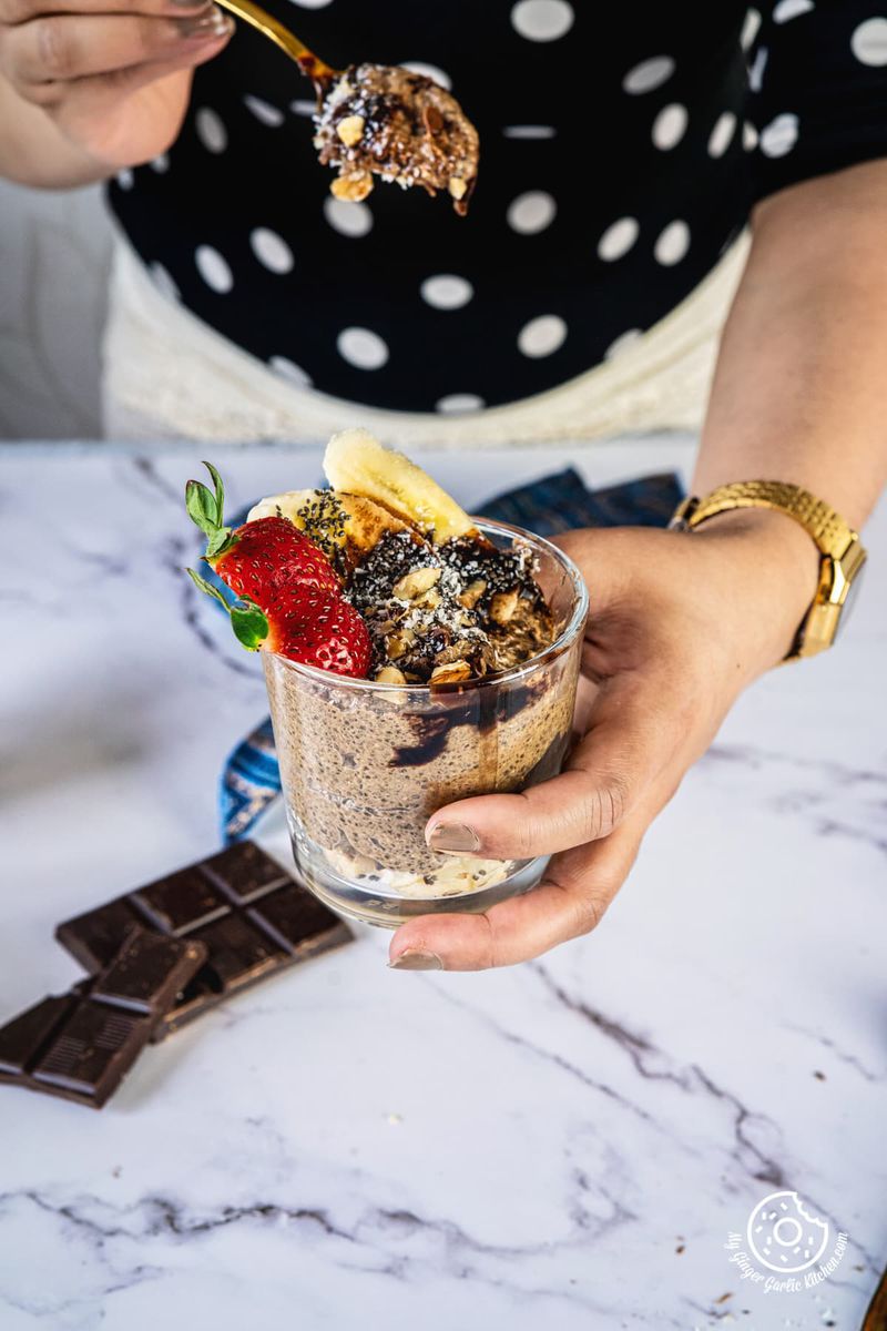 a hand holding chocolate coffee chia pudding glass in one hand and a golden spoon in other hand