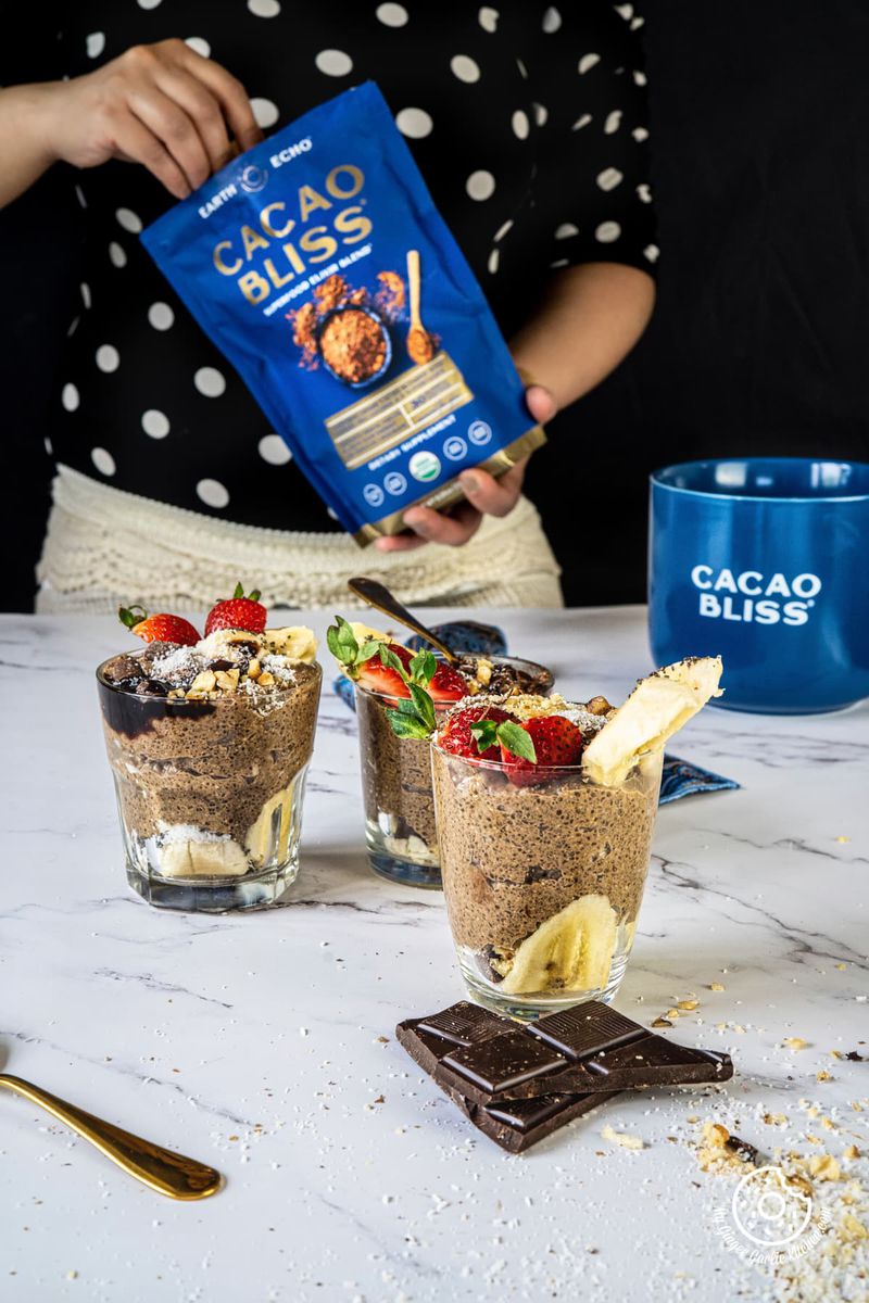 three chocolate coffee chia pudding glasses kept on a white backdrop and a female holding a chocolate powder pouch in the background