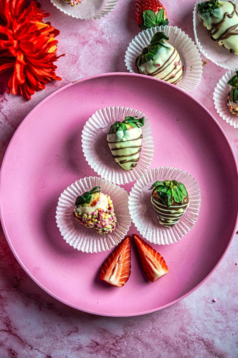 Chocolate covered strawberries with decorative drizzle in paper cups on a pink plate beside a red flower