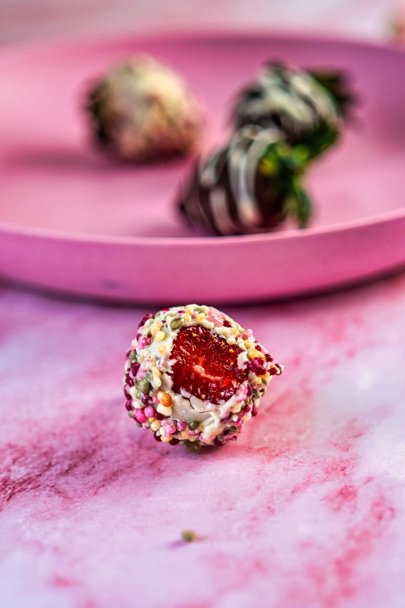 A bitten strawberry covered in white chocolate and colorful sprinkles on a pink plate