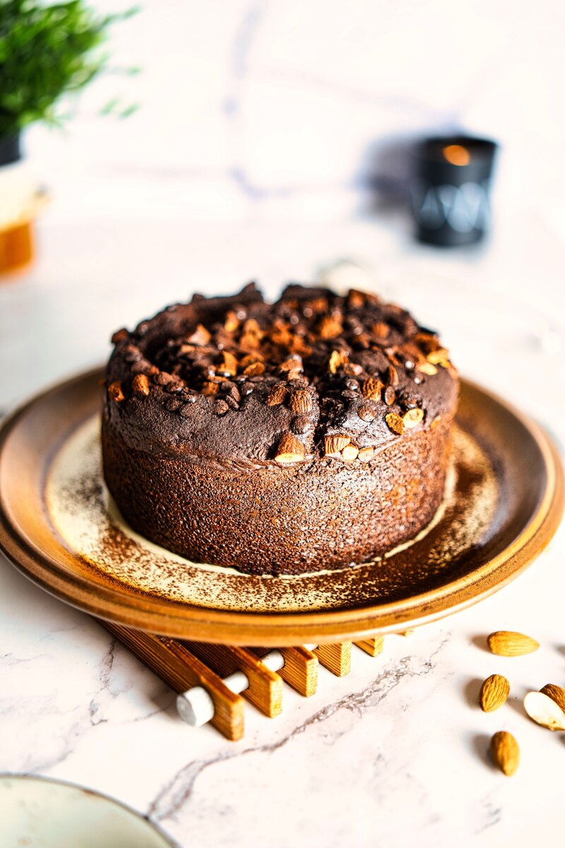 Whole vegan chocolate date cake on ceramic plate, cocoa-dusted surface with crunchy almonds and chocolate chips, easy eggless dessert for tea time