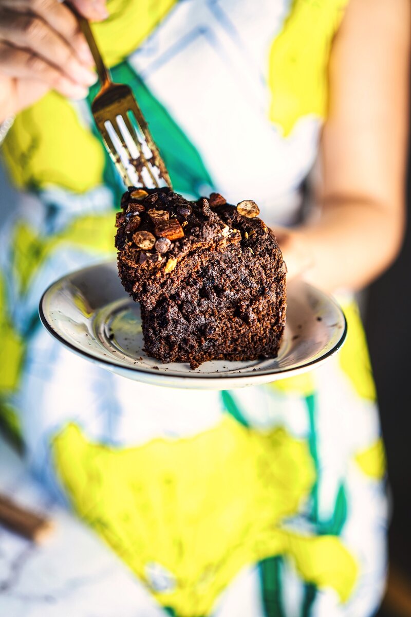 Hand holding slice of fudgy chocolate date cake on small plate, close-up showing rich texture with almonds and dark chocolate chips, healthy no-refined-sugar bake