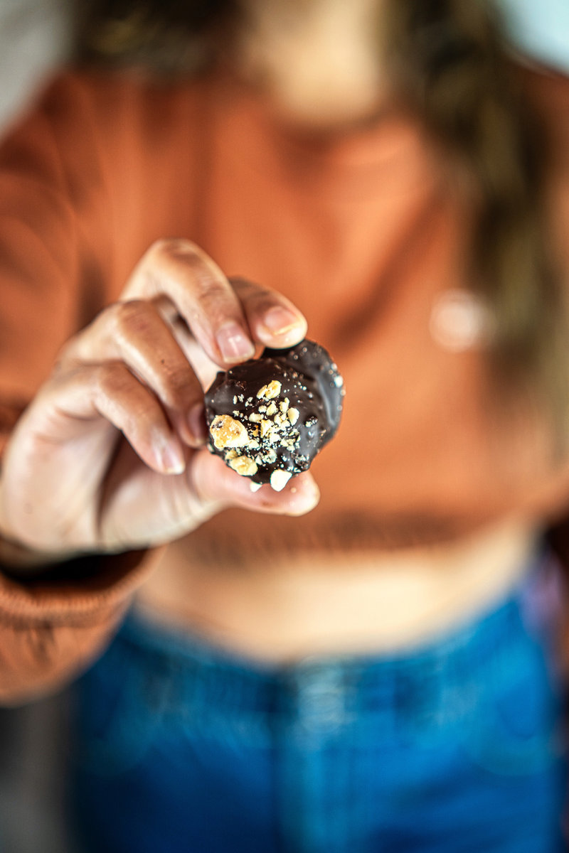 A person holding a chocolate hazelnut bliss ball topped with chopped nuts against a blurred background.