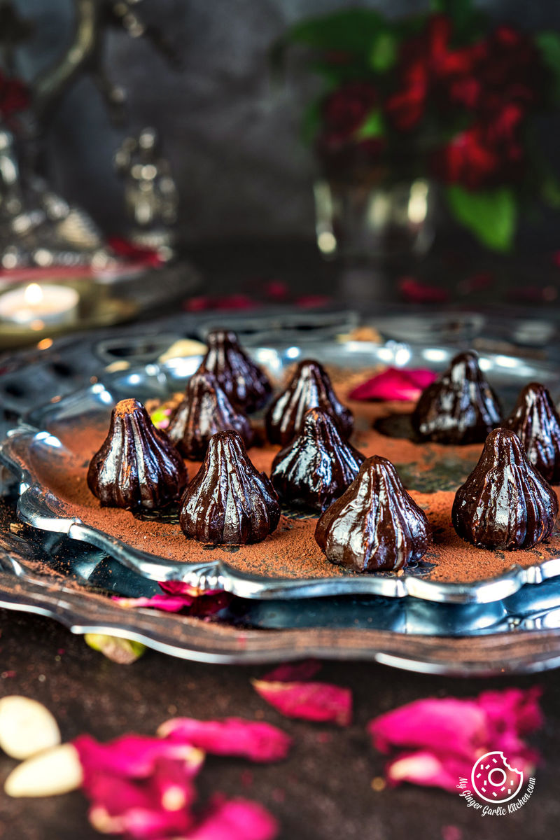chocolate modak on a steel plate garnished with dried rose petals with flowers in the background
