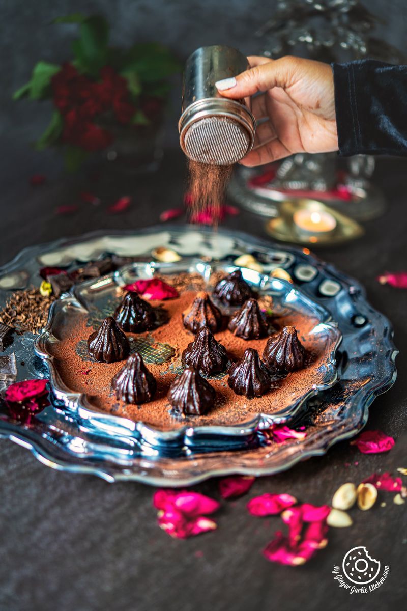 chocolate modak on a steel plate garnished with dried rose petals with a female sprinkling cocoa powder on modaks