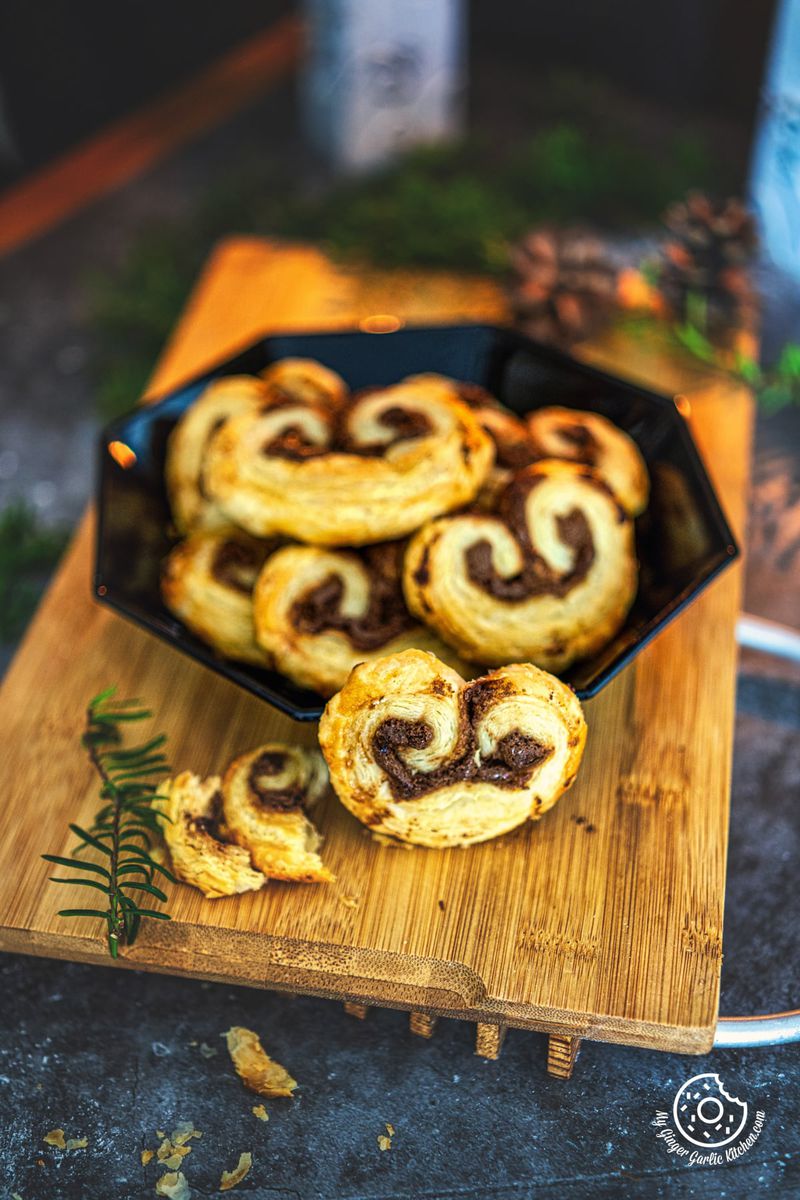 one chocolate palmier on a wooden board with a black plate stacked with chocolate palmiers