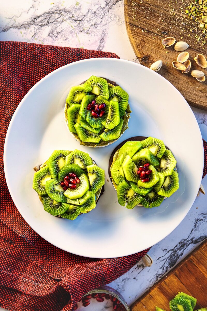 Close up of gluten free rice cake with dark chocolate base, pistachio butter topping and kiwi flower with pomegranate center