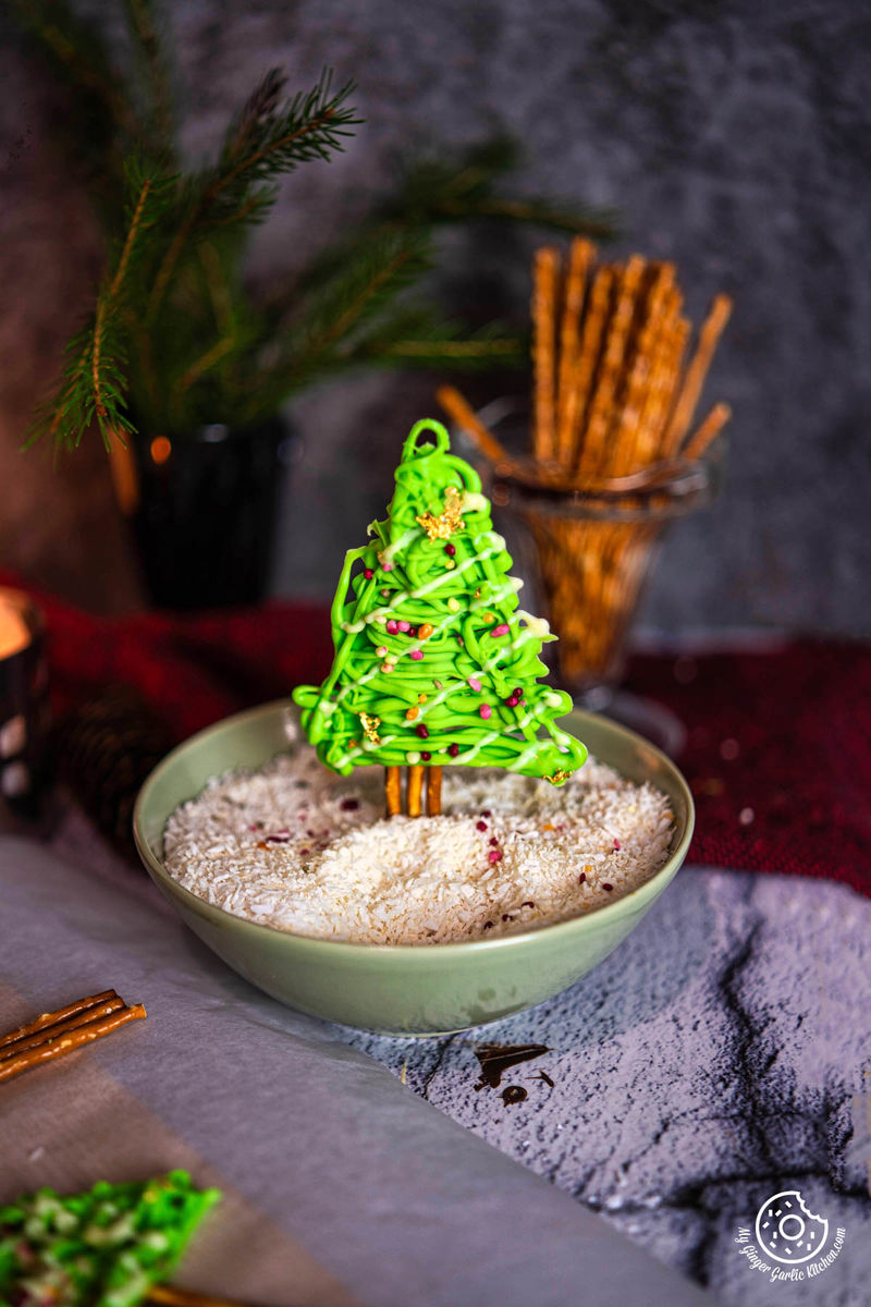 A chocolate pretzel Christmas tree sits in a bowl of coconut flakes. The tree is decorated with green and white chocolate and gold sprinkles.