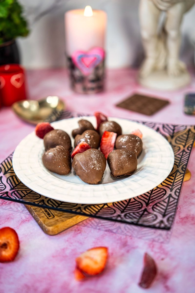 Heart-shaped chocolate covered strawberries arranged on a white plate with romantic Valentine's Day décor including a lit candle in the background