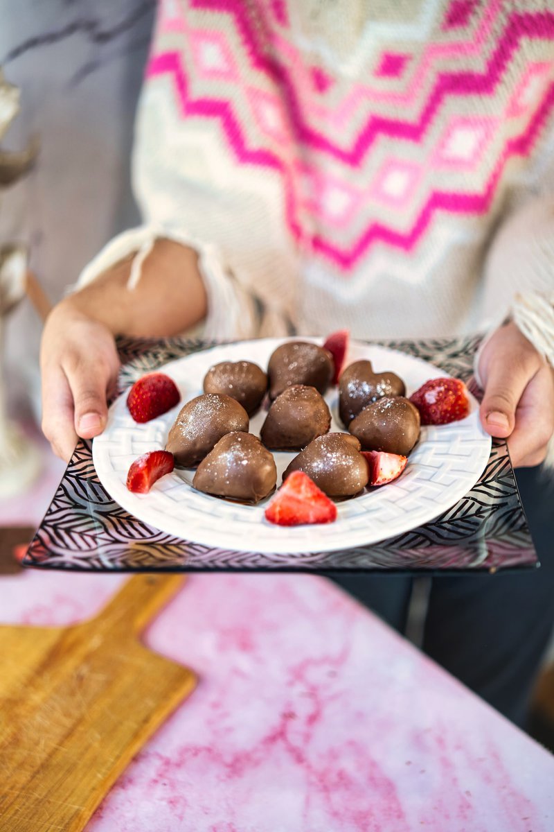 A person presenting a plate of chocolate-covered strawberry hearts with sliced strawberries on a patterned tray.