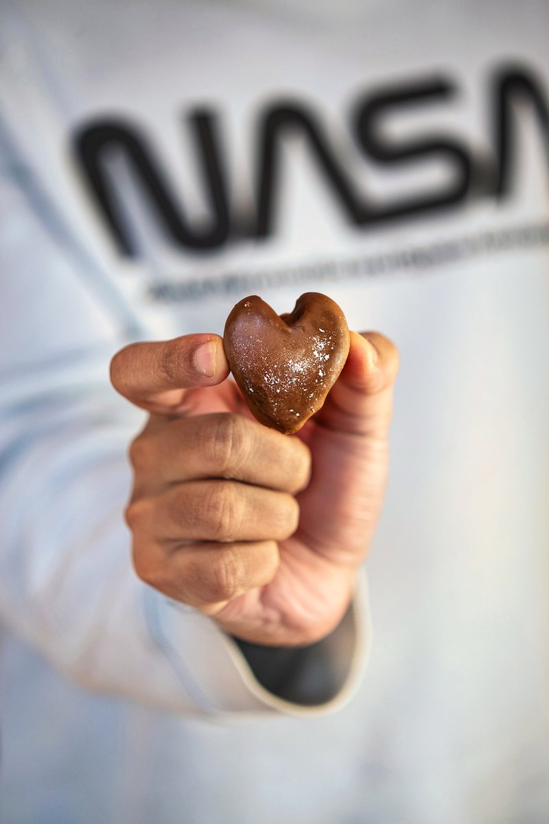 Close-up of a hand holding a single heart-shaped chocolate covered strawberry dusted with powdered sugar against a NASA logo background