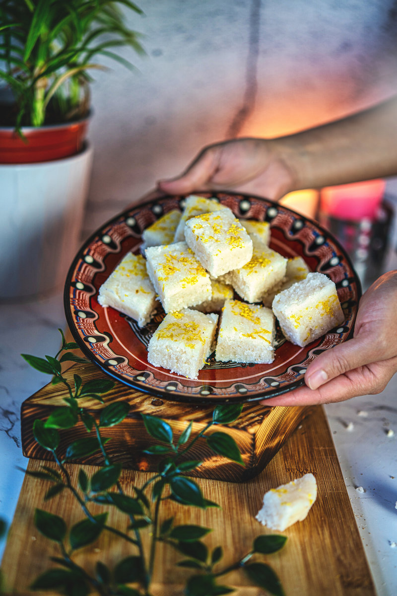 A person holding a plate of no-bake coconut bars garnished with lemon zest, presented on a rustic wooden board with a plant in the background.