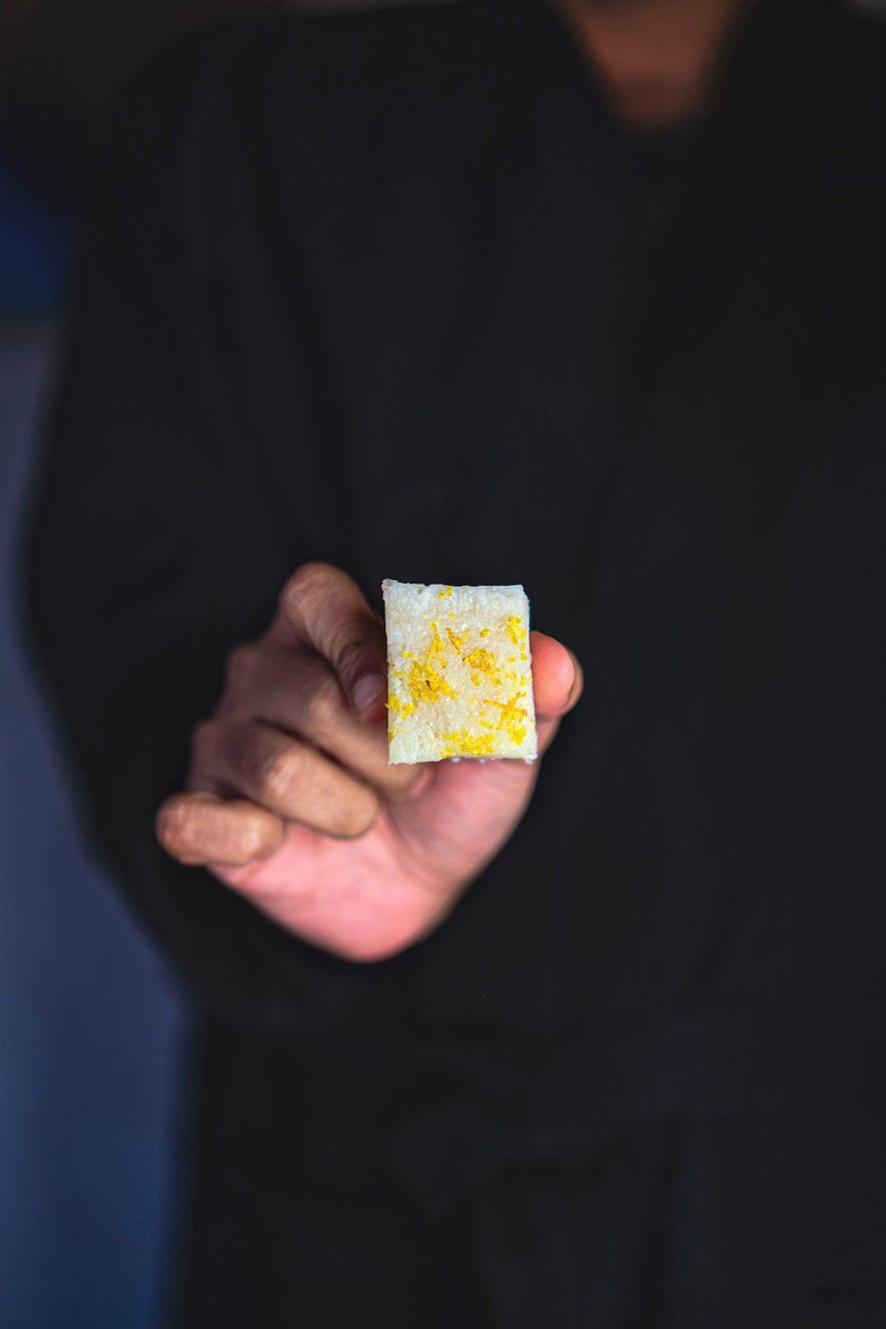 A hand holding a small square of coconut bar dessert with flecks of yellow, likely lemon zest, visible on its white surface.