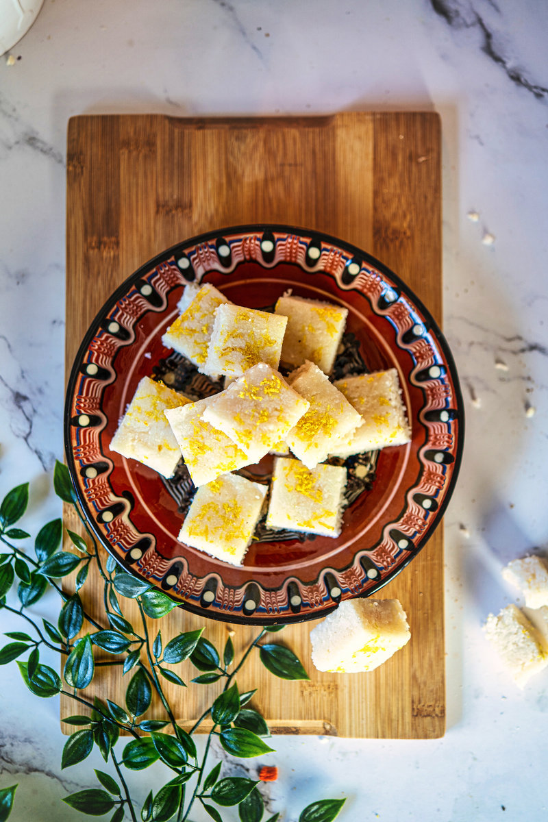 Top view of no-bake coconut bars with lemon zest served on a decorative red plate, placed on a wooden board with greenery for garnish