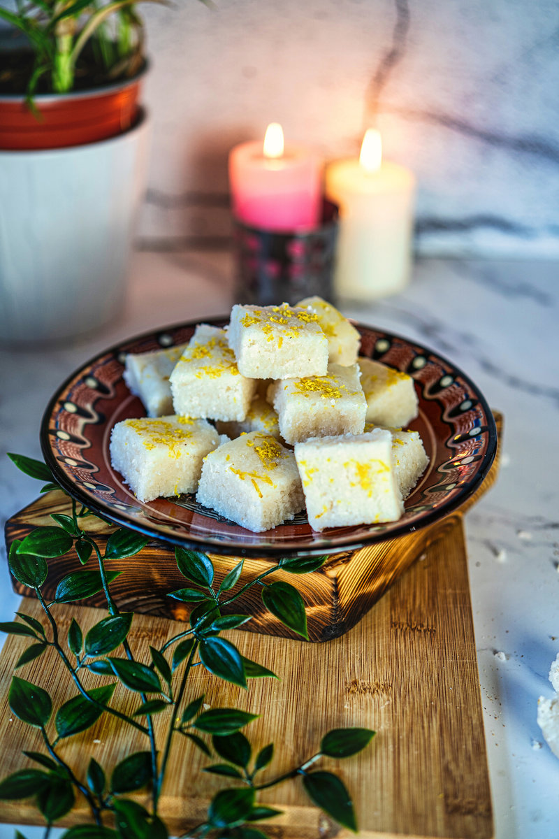 No-bake coconut bars with lemon zest arranged on a decorative plate, accompanied by candles and potted plants in the background.