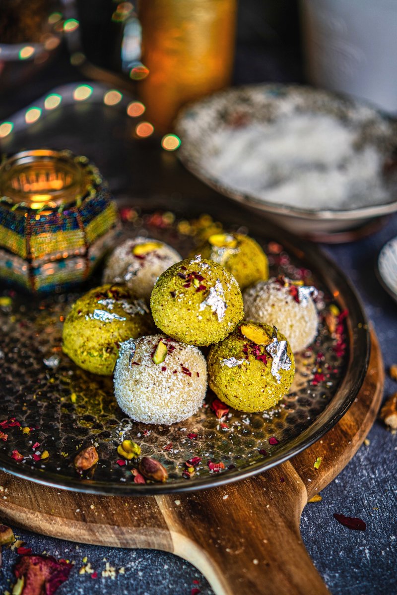 Coconut ladoo truffles with pistachio and coconut coatings, topped with edible silver leaf and pistachios on a festive serving plate.