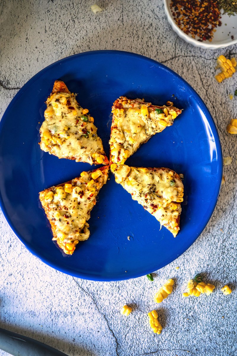 Four pieces of Corn Chilli Cheese Toast arranged on a blue plate, with scattered corn and seasoning.