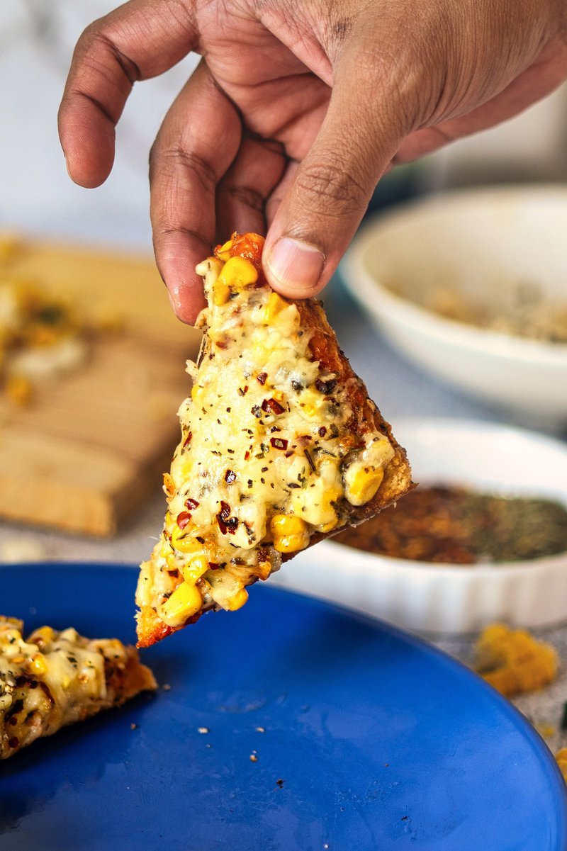 A hand holding a slice of Corn Chilli Cheese Toast, displaying the melted cheese and toppings.