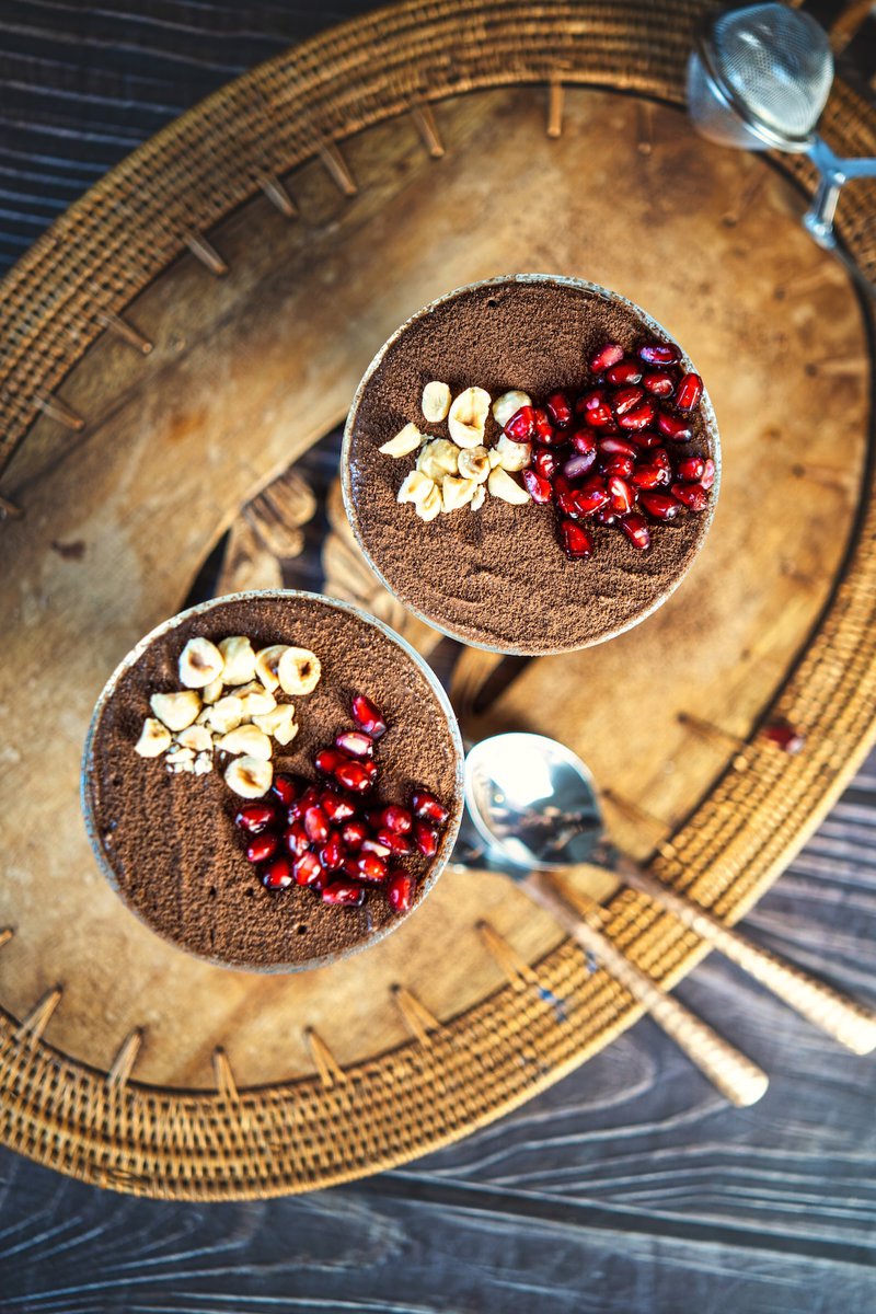 Top-down view of two glasses of cottage cheese chocolate mousse, garnished with chopped nuts and pomegranate seeds on a woven placemat.