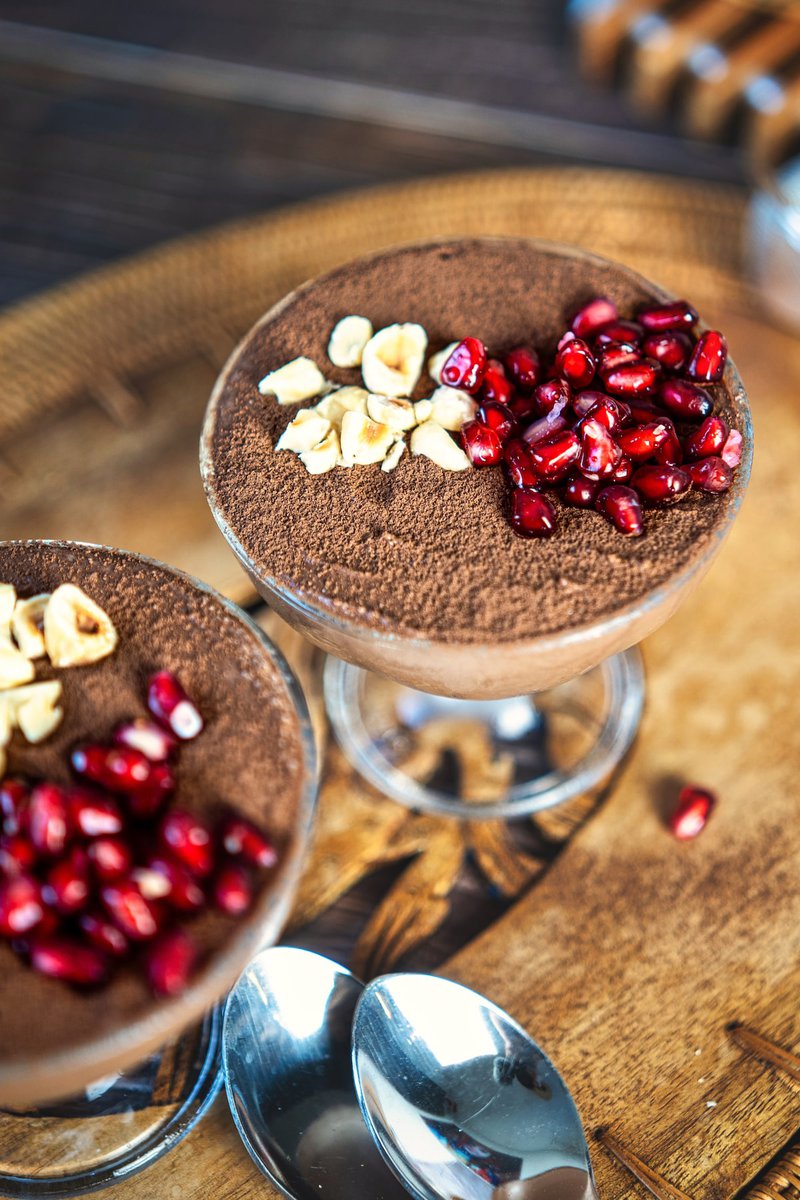 Two glass bowls of chocolate mousse garnished with chopped nuts and pomegranate seeds, accompanied by two spoons on a wooden tray.