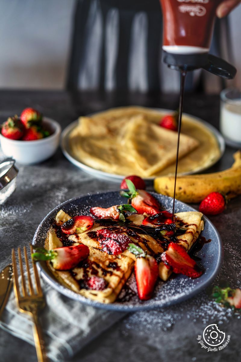 chocolate sauce drizzling over a gray plate with two folded sweet crepes topped with strawberries and chocolate sauce