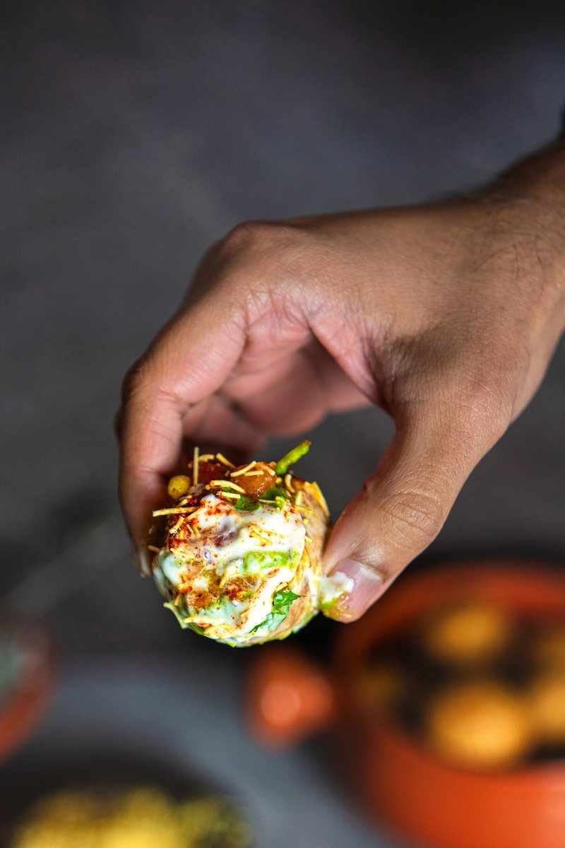 A hand holding a single Dahi Puri, topped with yogurt, chutneys, and spices, with a blurred background