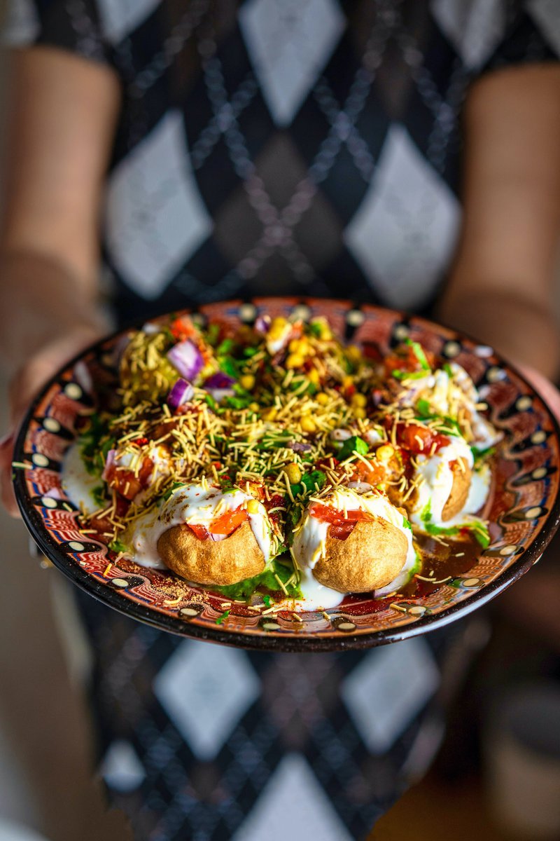 A person holding a plate of Dahi Puris, richly garnished with yogurt, chutneys, and spices, with a focus on the colorful toppings.