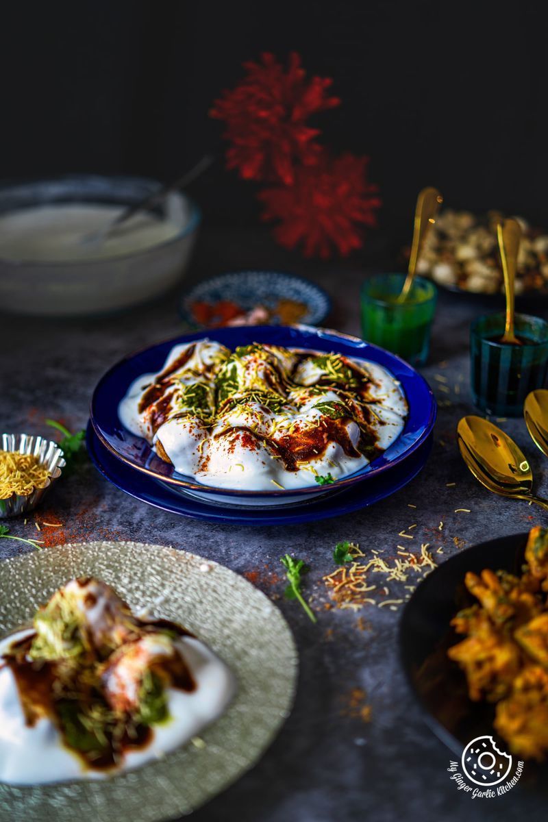 A beautifully presented blue plate of dahi vada garnished with spices and chutney, with bowls of sev, chutney and spices in the background, on a dark textured surface.