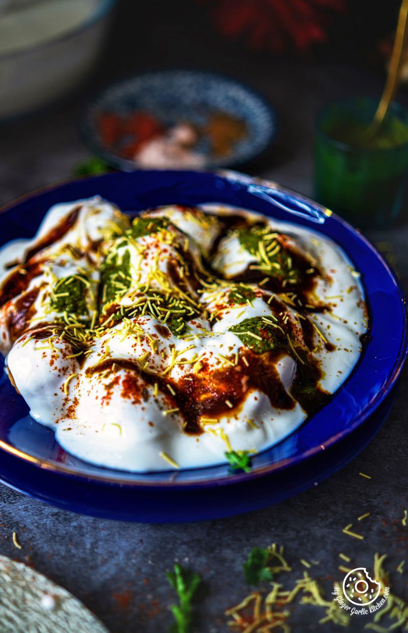a closeup shot of dahi vada topped with chutneys and sev in a blue glass plate, with dark moody lighting.