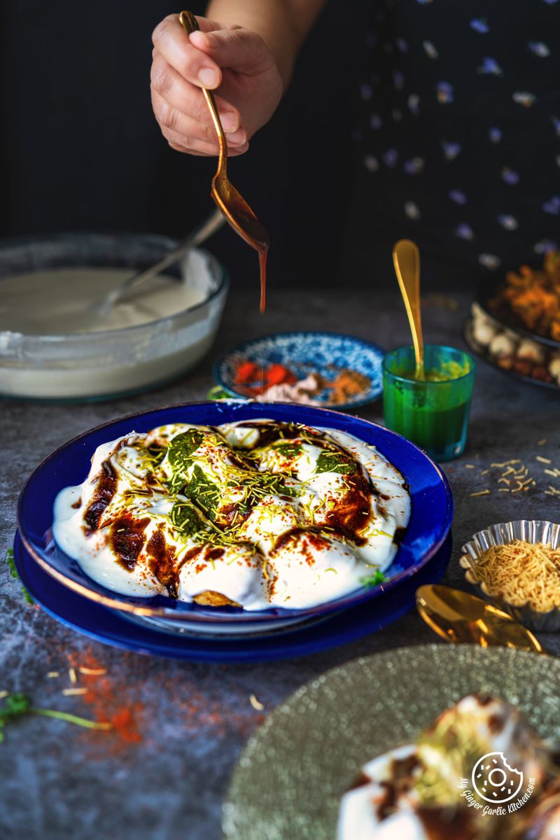 a hand pouring tamarind chutney over a dahi vada plate topped with chutneys and sev, with kitchen utensils in the background