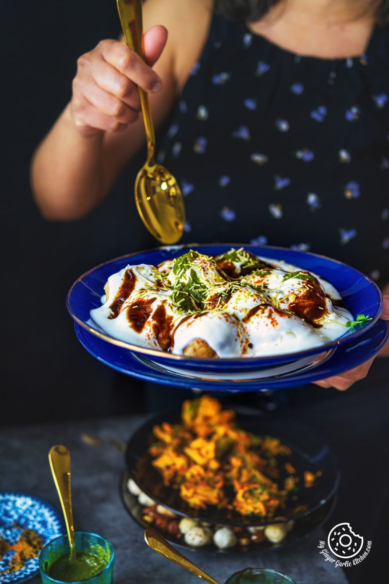 a female holding a golden spoon in one hand and blue glass plate filled dahi vada aka dahi bhalla garnished with chutneys and sev with other hand