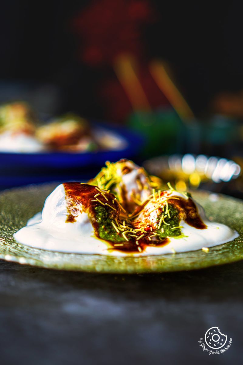 A single serving of dahi vada on a shimmering plate, drizzled with green and tamarind chutneys, and garnished with fine sev, highlighted by a moody ambiance.