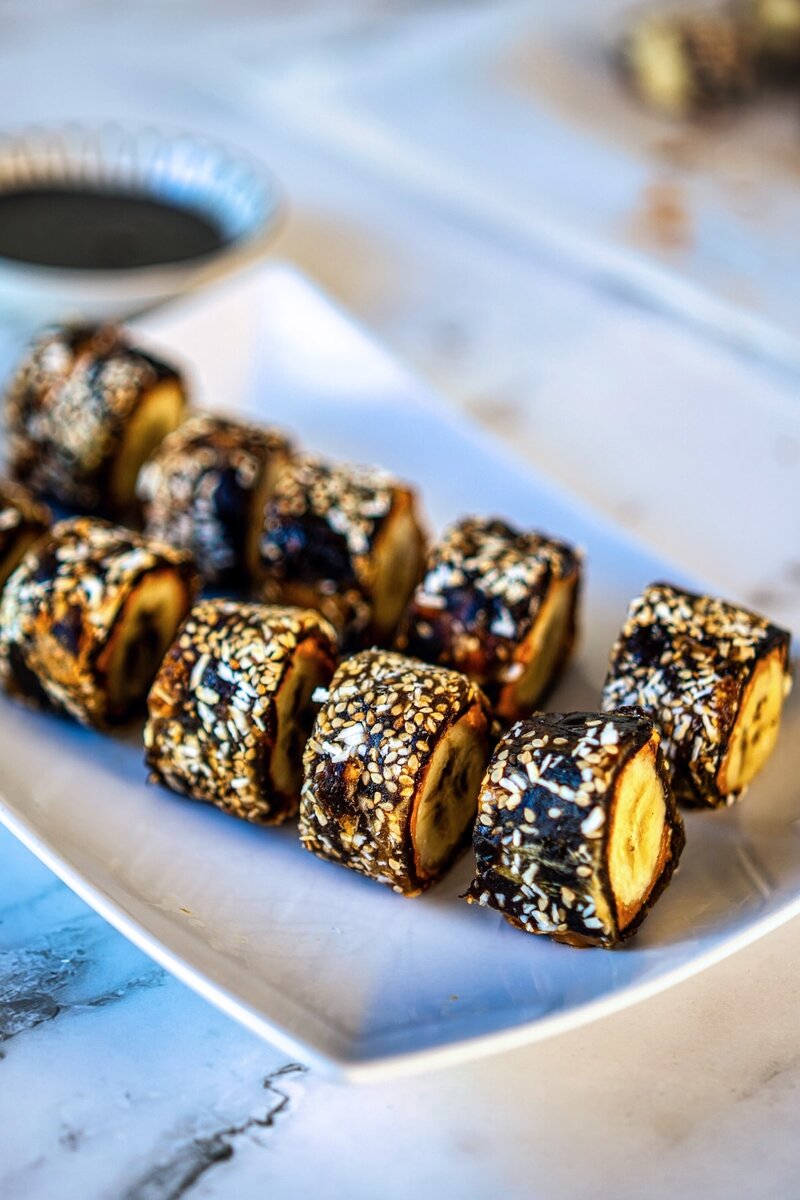 Date banana sushi sliced and arranged on a white plate with sesame seeds and coconut coating