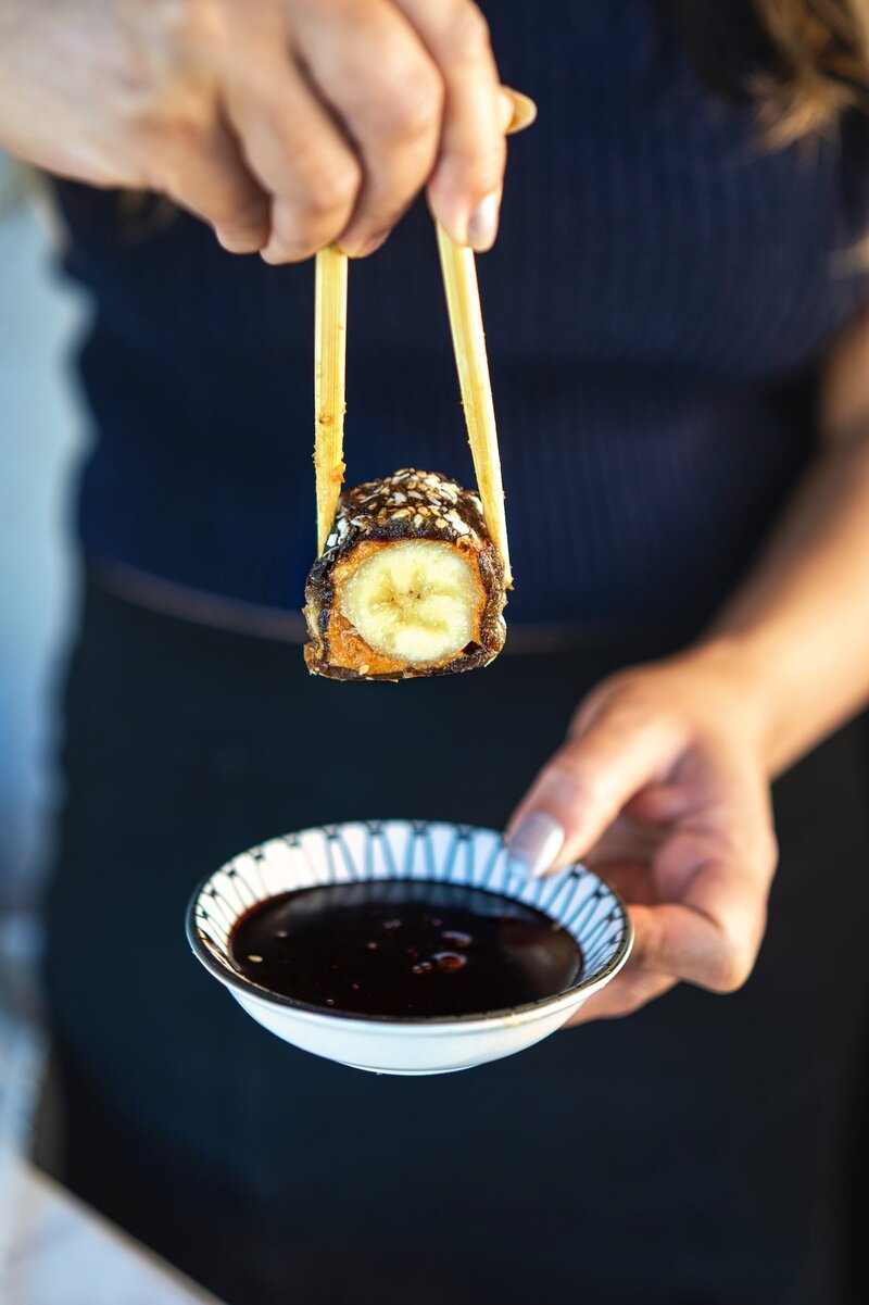 Close up of date banana sushi held with chopsticks dipped into chocolate sauce