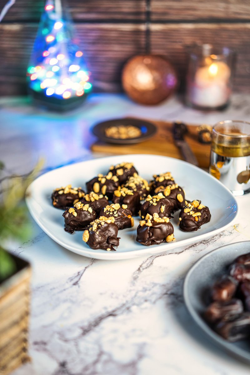 A plate of homemade date Snickers coated in dark chocolate and crushed peanuts, on a plate with festive lights in the background.