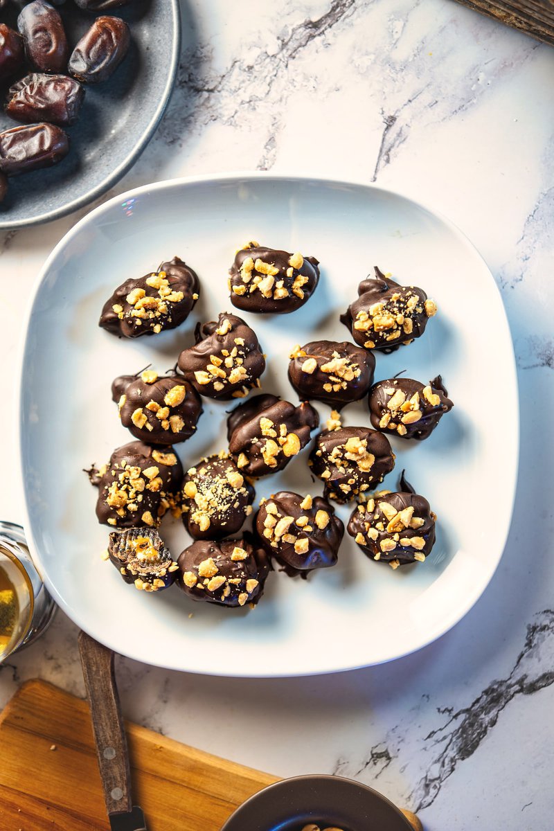  Plate of chocolate-covered dates topped with crushed nuts on a marble surface.