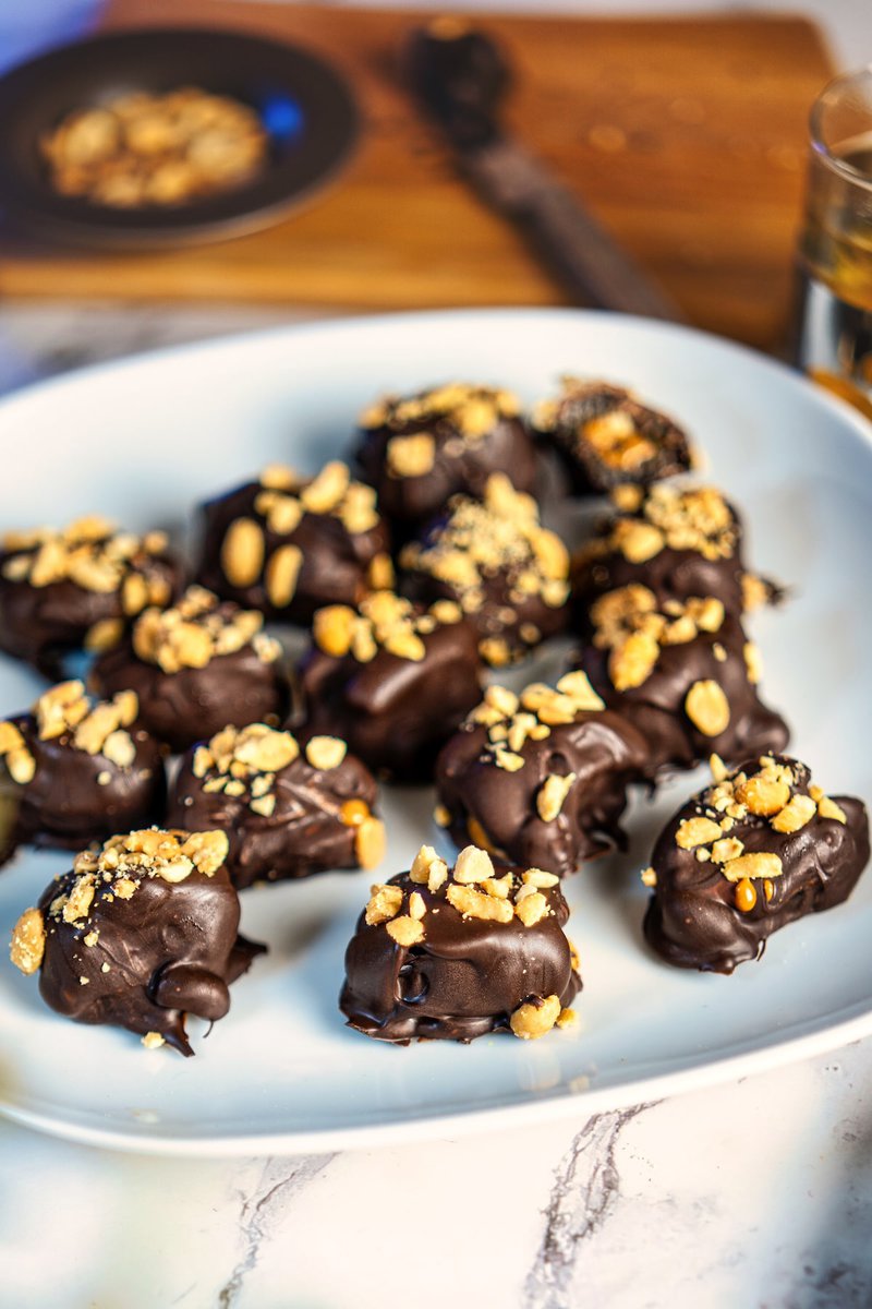 Close-up view of dark chocolate-coated date Snickers garnished with crushed peanuts, arranged on a white plate, perfect as a healthy snack or dessert.