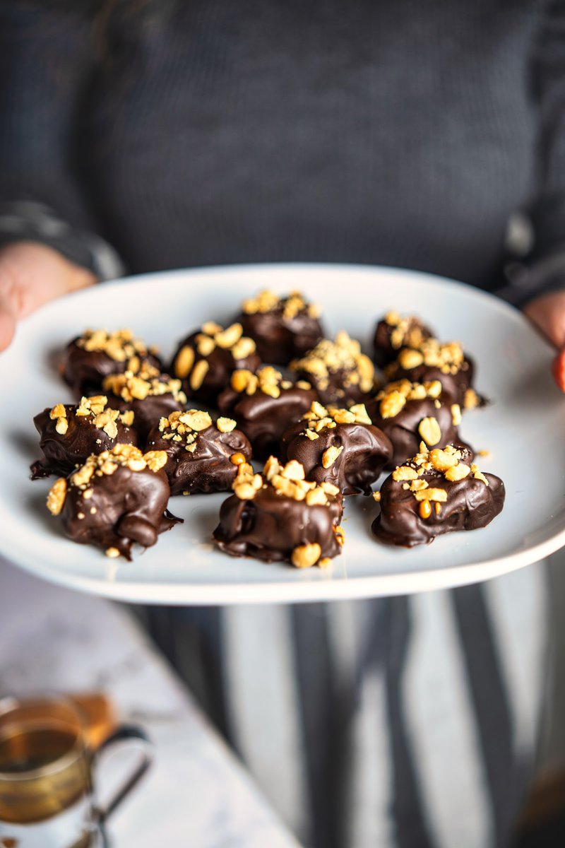 A person holding a white plate filled with decadent chocolate-covered date Snickers topped with crushed roasted peanuts, ideal for gifting or enjoying at festive gatherings.