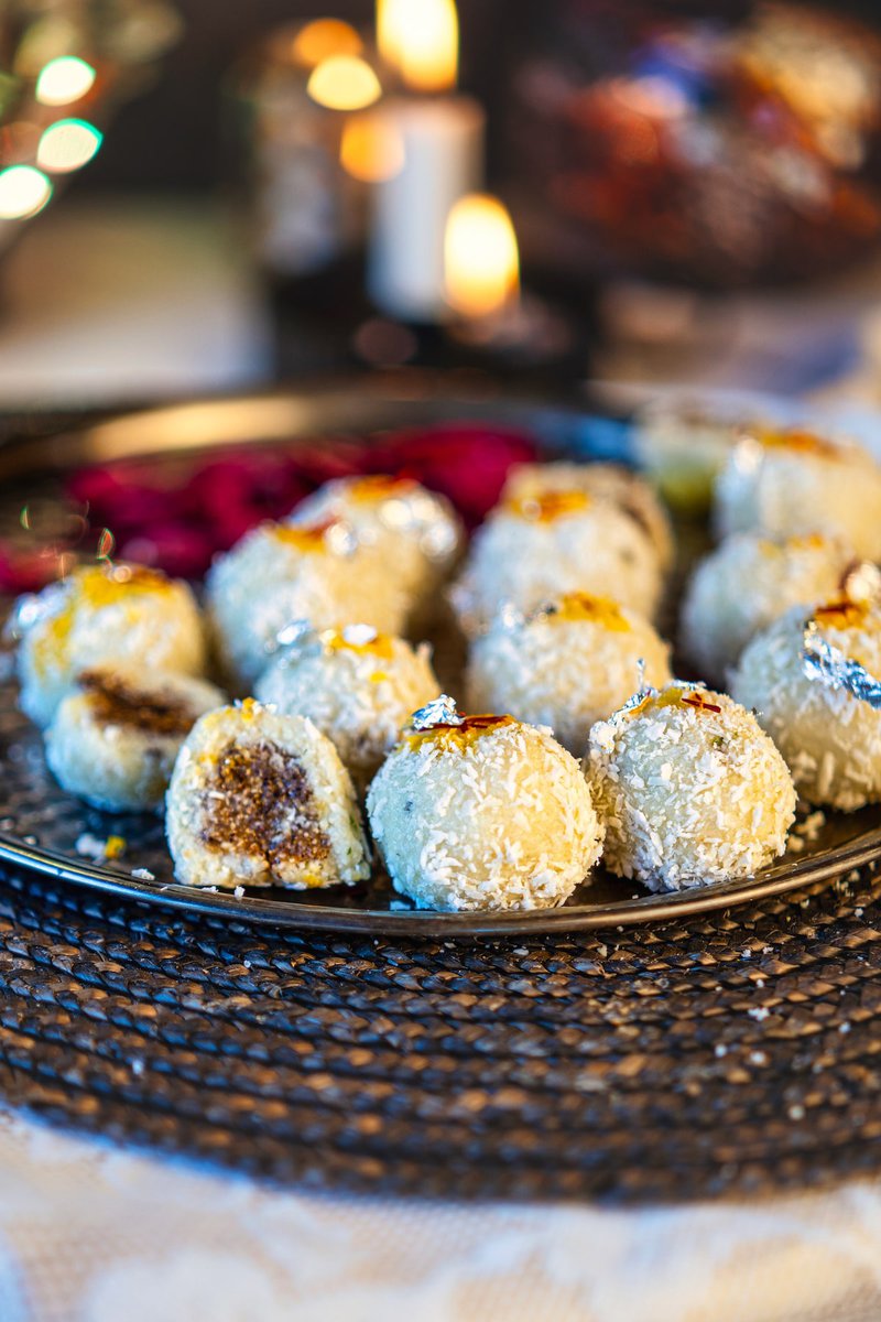 Plate of date-stuffed coconut ladoos garnished with edible silver leaves, surrounded by festive lights.