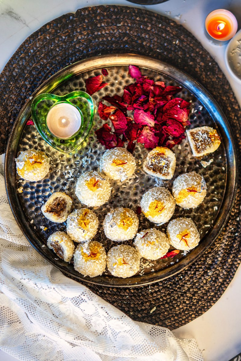 A tray of date-stuffed coconut ladoos garnished with saffron, surrounded by rose petals and a small decorative candle.