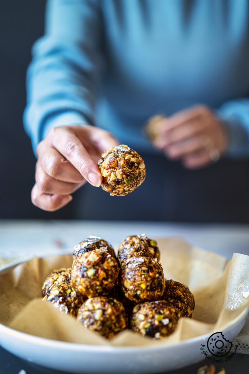 a female holding a dry fruit laddu over a white plate with filled with laddus