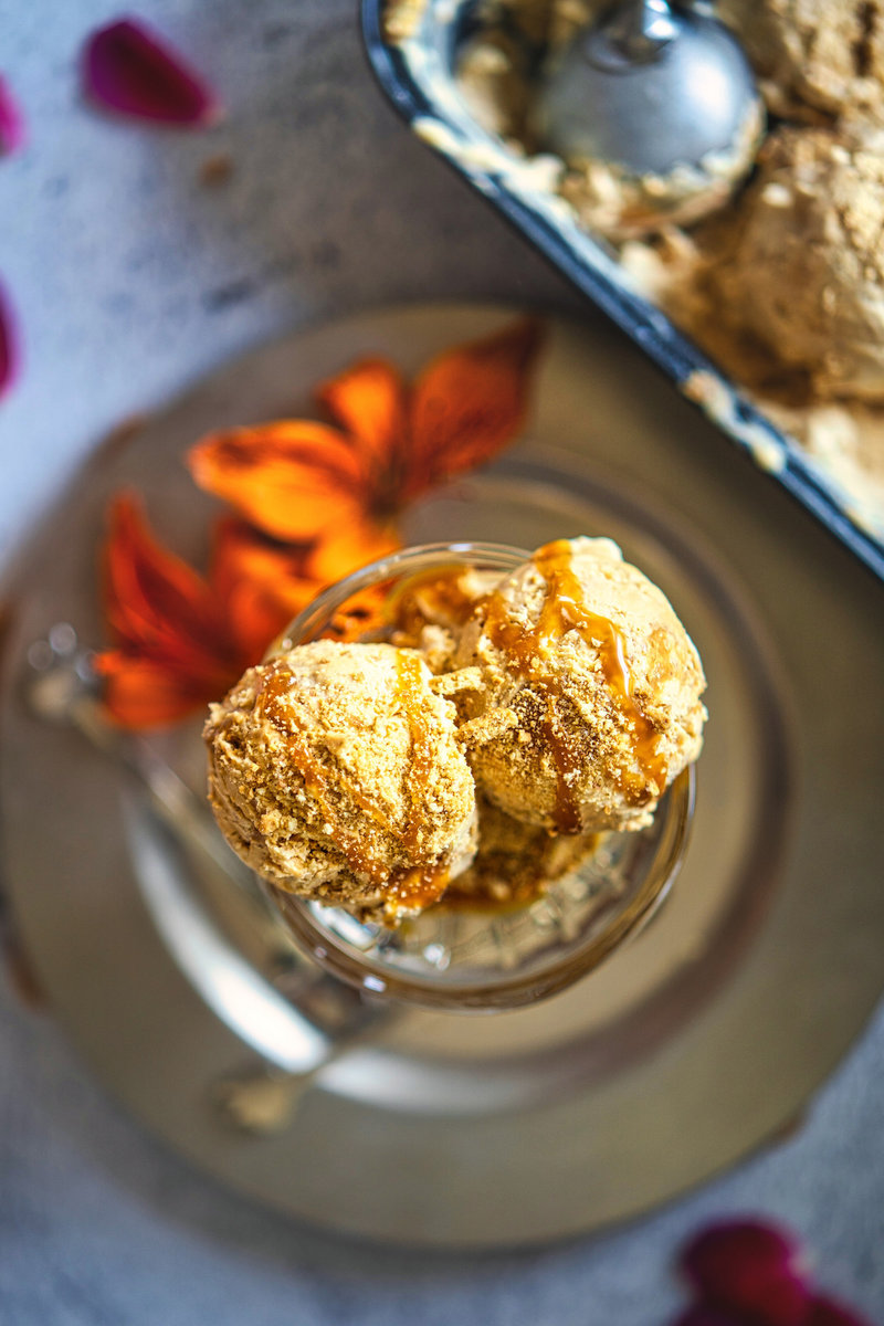 Two scoops of dulce de leche ice cream in a glass dish, drizzled with caramel and topped with crushed cookies, on a plate with orange flowers