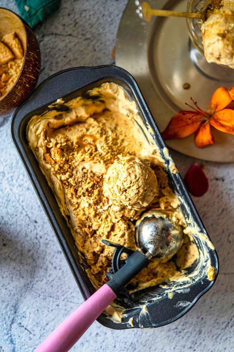 Metal container of dulce de leche ice cream with a scoop and cookie crumbles, alongside a pink-handled scoop and a flower