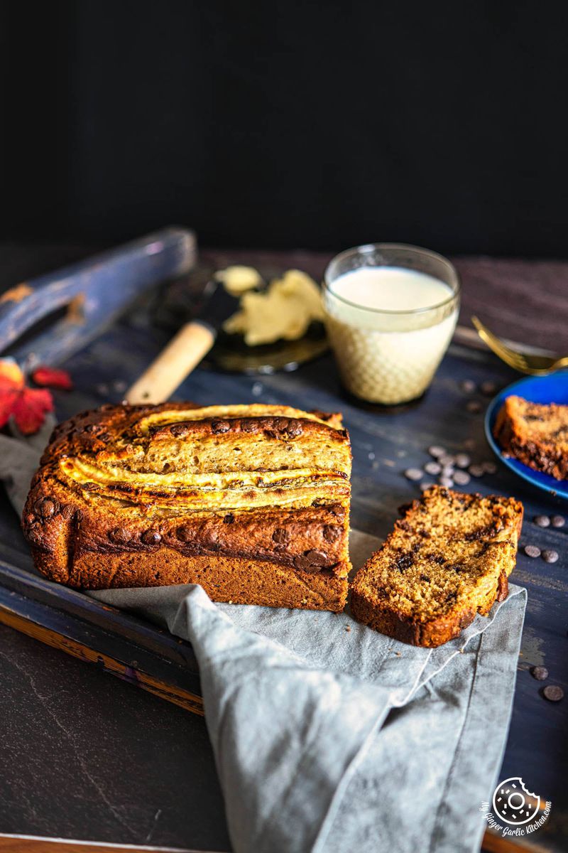 a slice of whole wheat eggless banana bread sits on a tray next to a banana bread loaf and a glass of milk