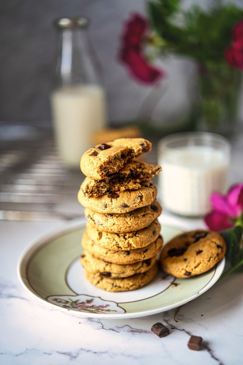 A stack of eggless chocolate chip cookies on a floral plate, with a glass of milk in the background.