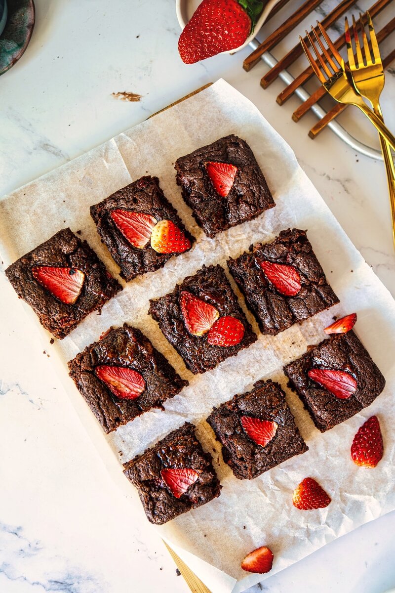 Nine chocolate brownies topped with sliced strawberries, arranged on parchment paper.