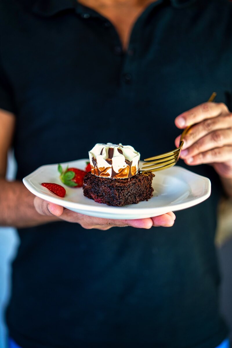 Person holding a plate with a chocolate brownie topped with whipped cream and chocolate drizzle, garnished with strawberries.