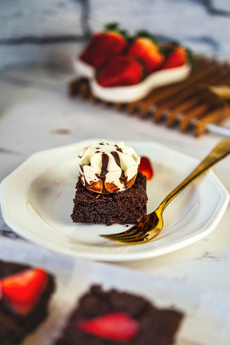 A single serving of chocolate brownie topped with vanilla ice cream and chocolate drizzle on a white plate with a gold fork, with fresh strawberries and additional brownies blurred in the background.