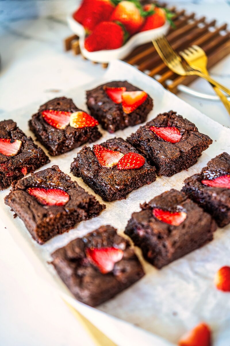 Rich chocolate brownies decorated with fresh strawberry slices displayed on parchment paper, with a small bowl of strawberries and golden utensils in the background.