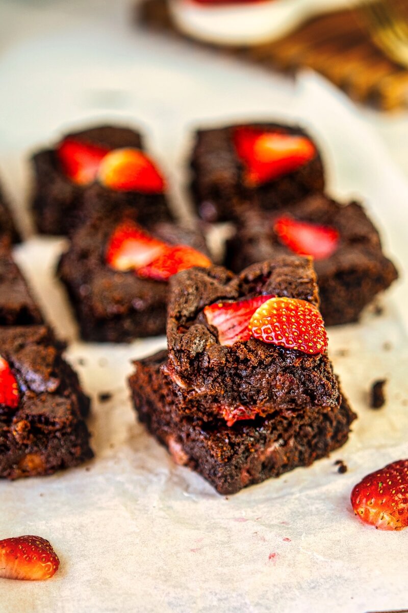 Stack of two chocolate brownies with strawberry slices, surrounded by more brownies on parchment paper.
